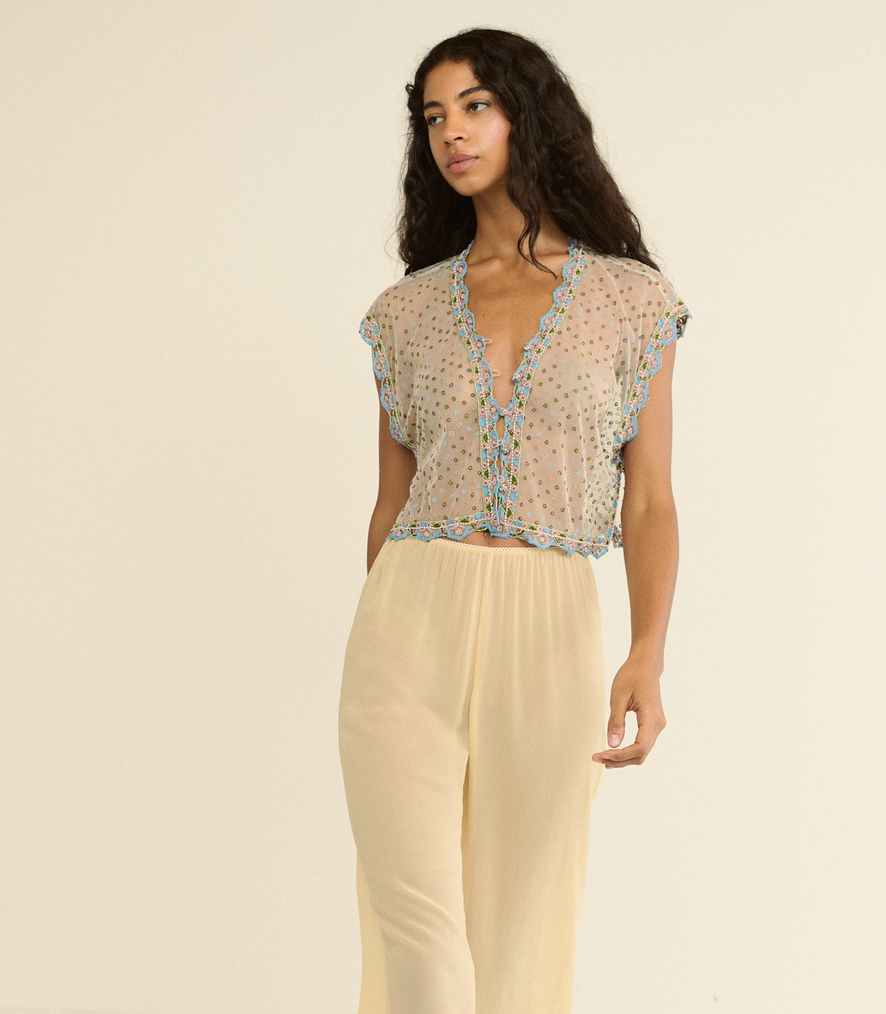 Woman wearing a sheer top with floral embellishments and light-colored pants on a beige background