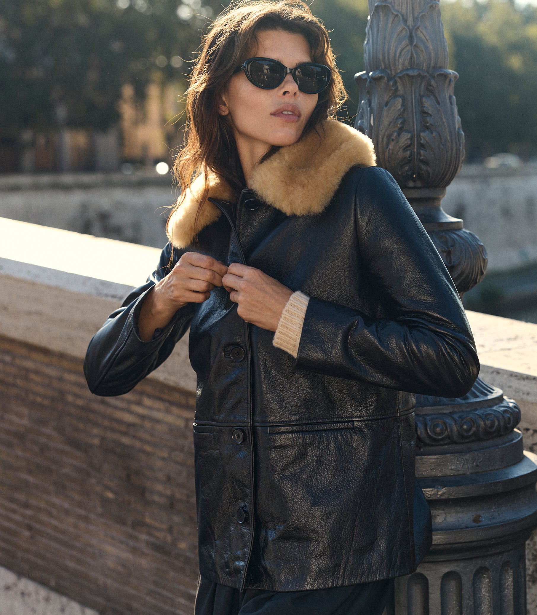 A woman wearing a black leather jacket with a beige fur collar and black sunglasses, posing outdoors next to a stone bridge and decorative metal post. 1
