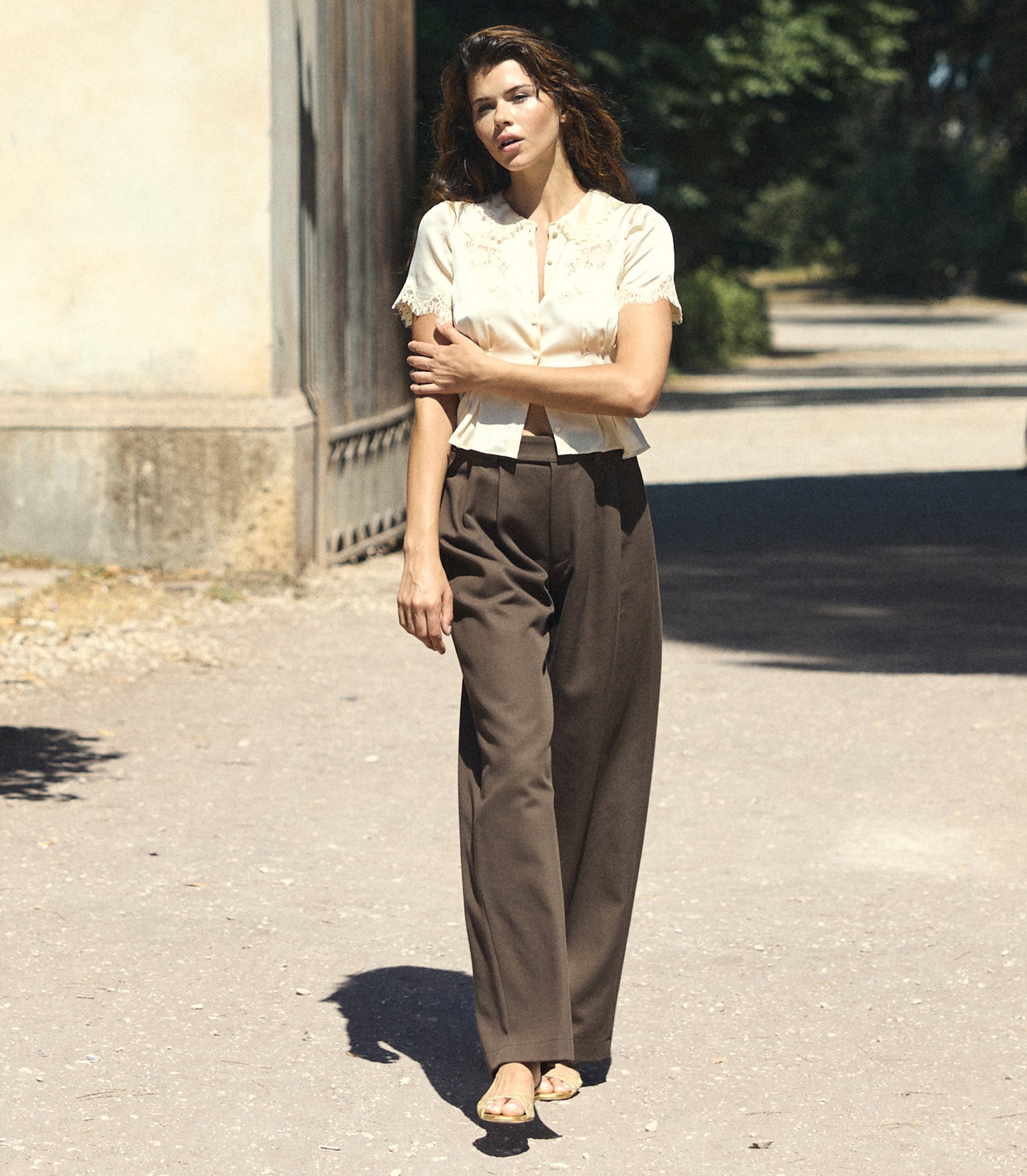 A woman stands outdoors on a sunlit paved path wearing a cream short-sleeve blouse with a ruffled collar and dark brown wide-leg trousers.