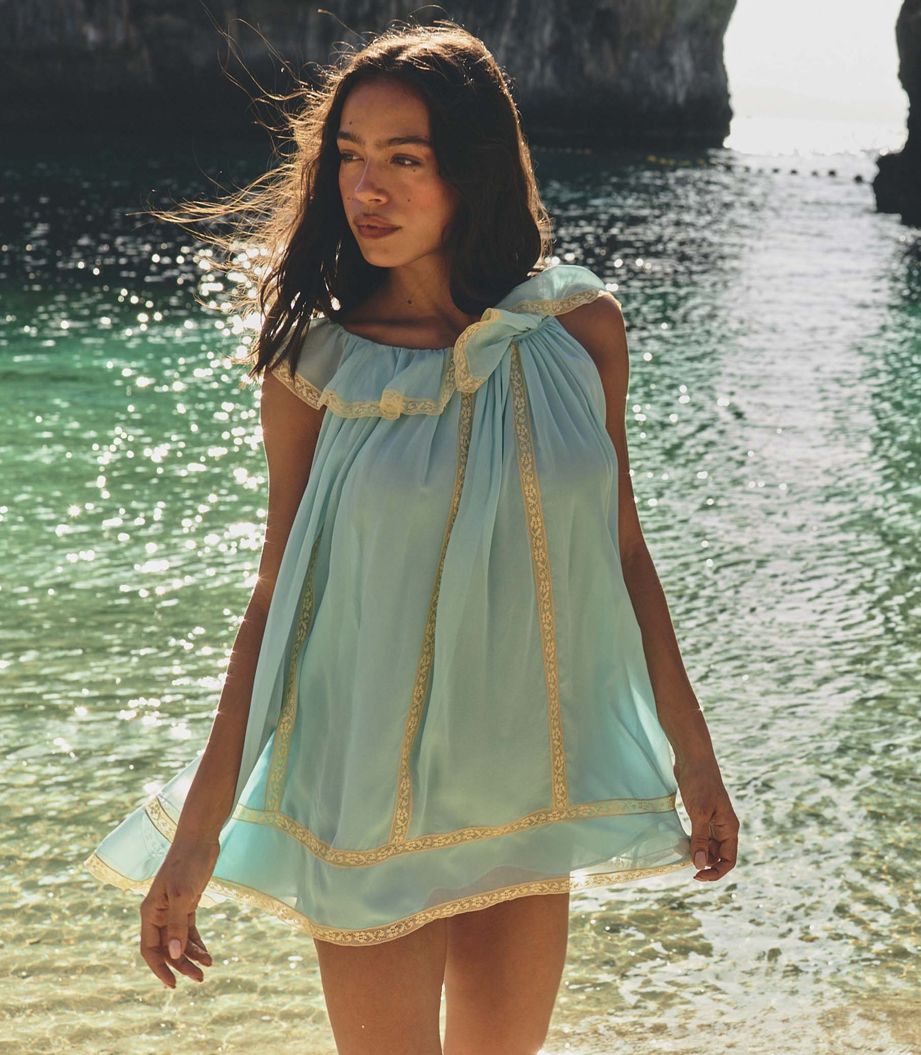 Woman in a light blue dress standing by a body of water with sunlight filtering through.