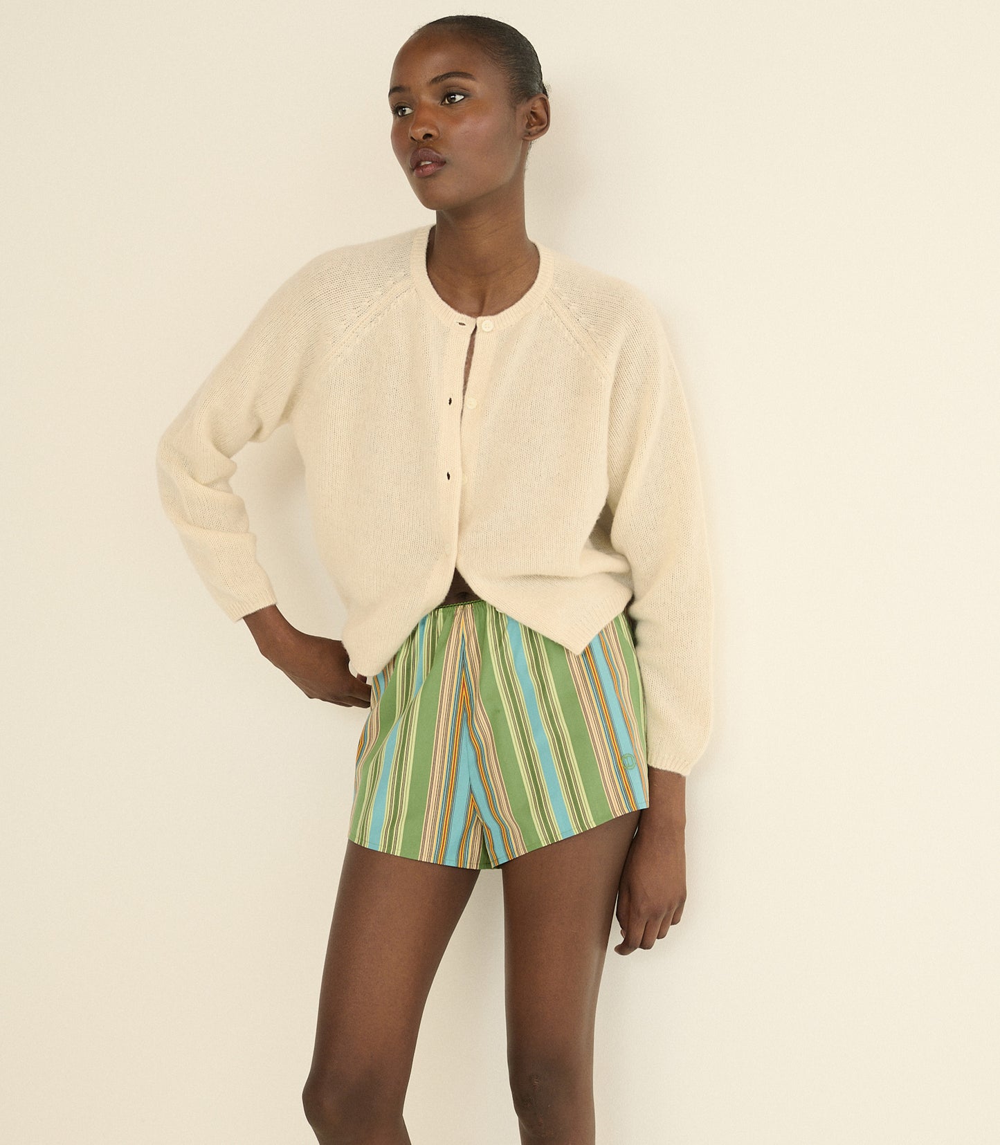 A model wearing a cream-colored knit cropped cardigan and green, blue, and orange striped shorts, standing against a plain off-white background.