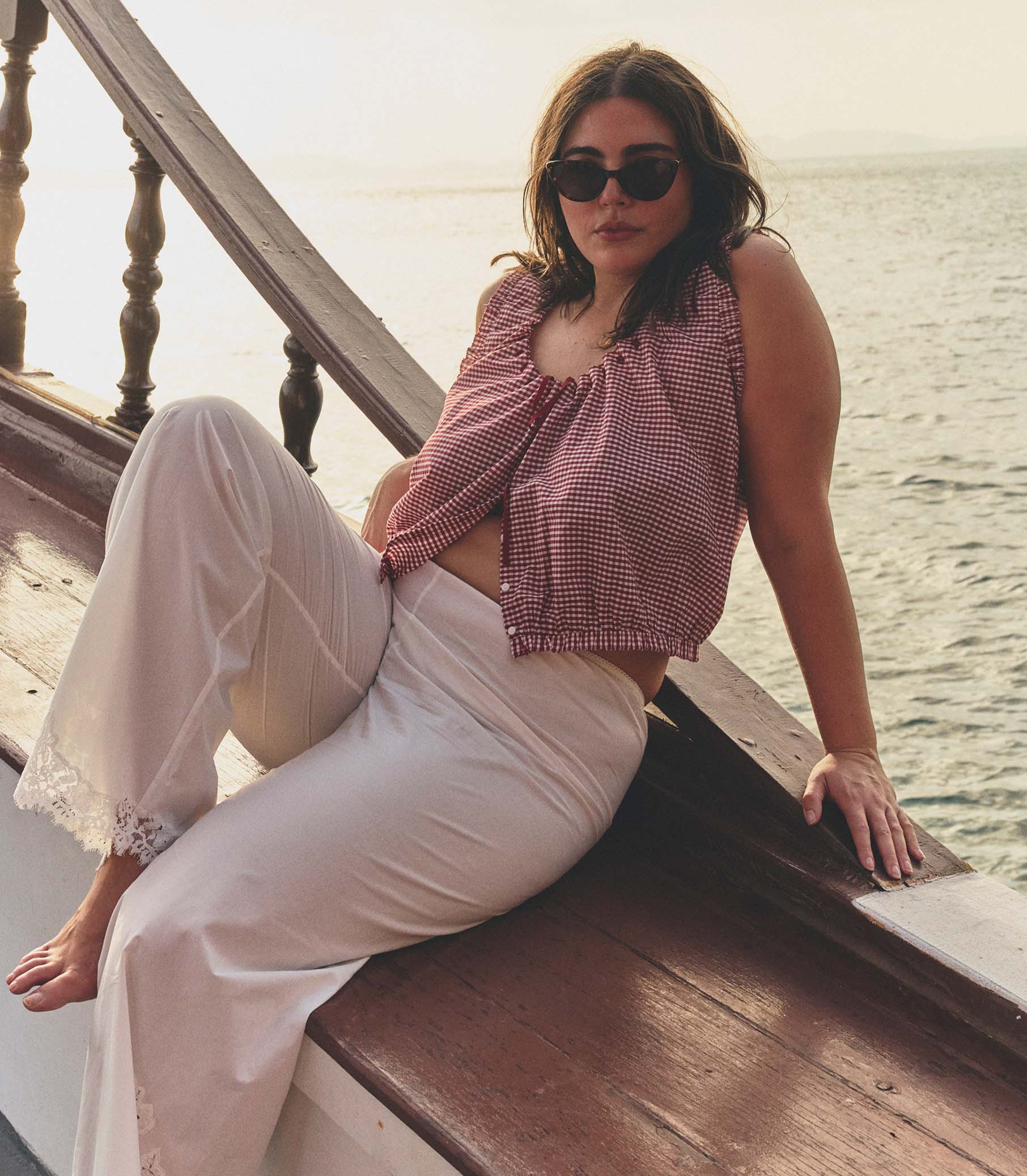 Woman sitting on a boat deck by the water wearing sunglasses and a red checkered top with white pants.