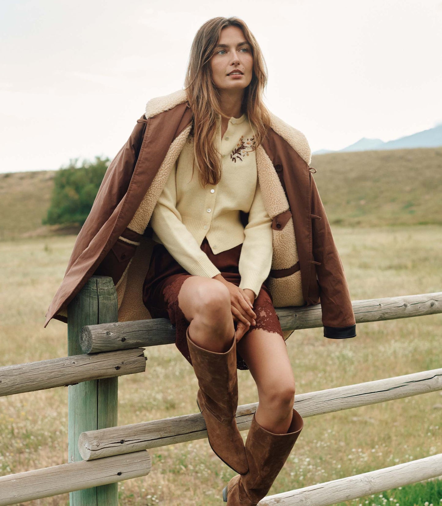 Woman sitting on a wooden fence in a field wearing a brown coat and knee-high boots.