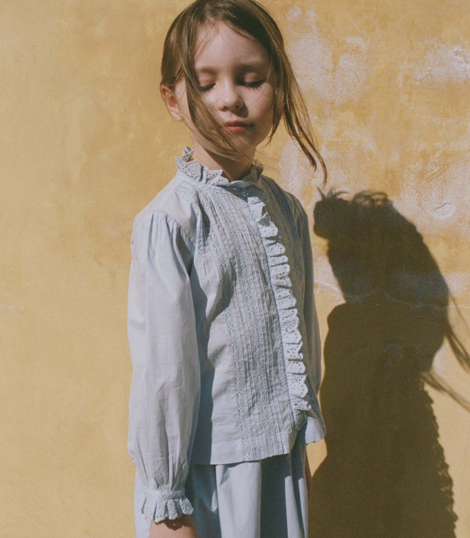 Young girl in a light blue blouse standing against a textured yellow wall with a soft focus. 1

