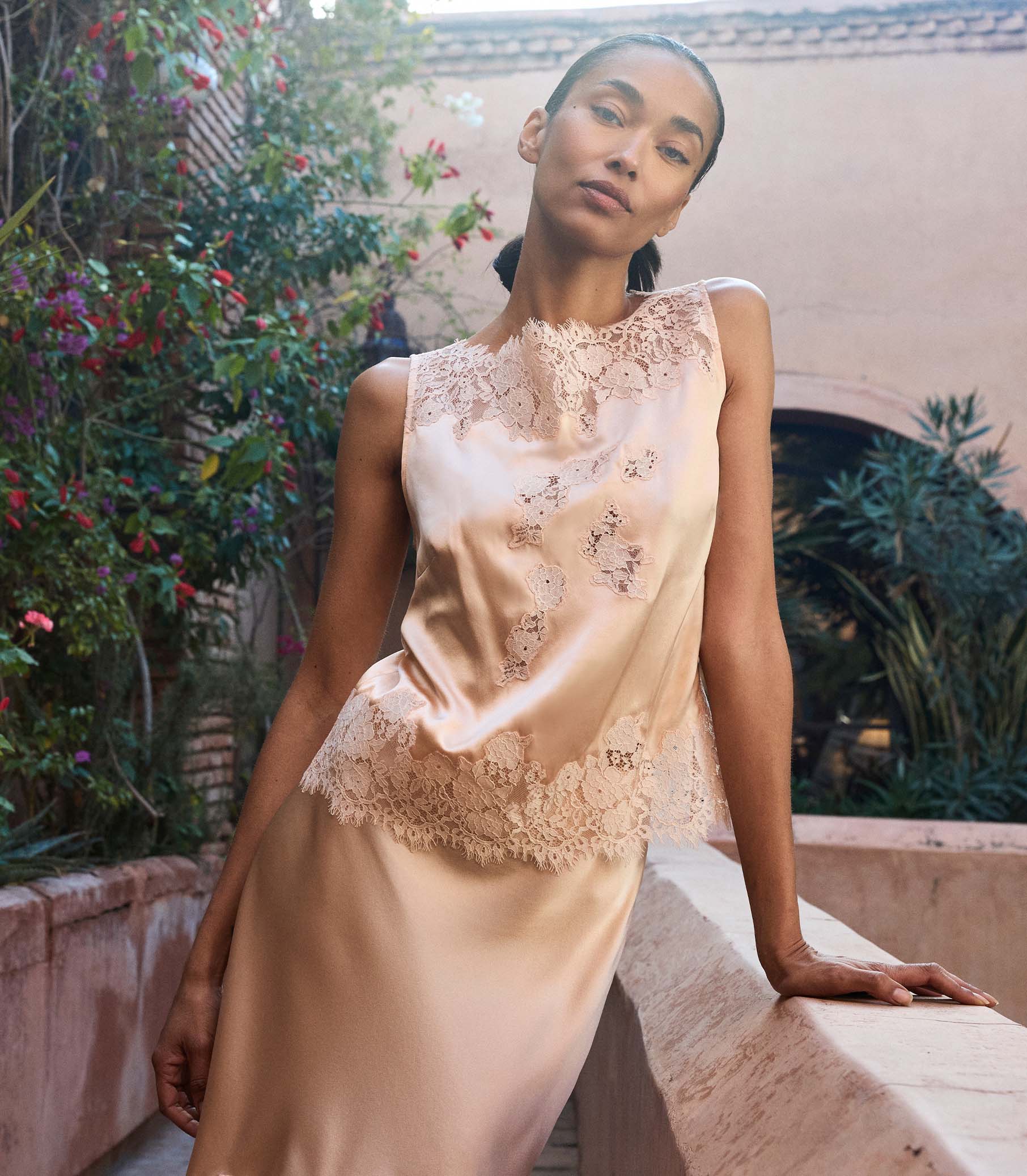 Woman wearing a peach lace blouse with a matching skirt sitting outdoors with greenery in the background 1
