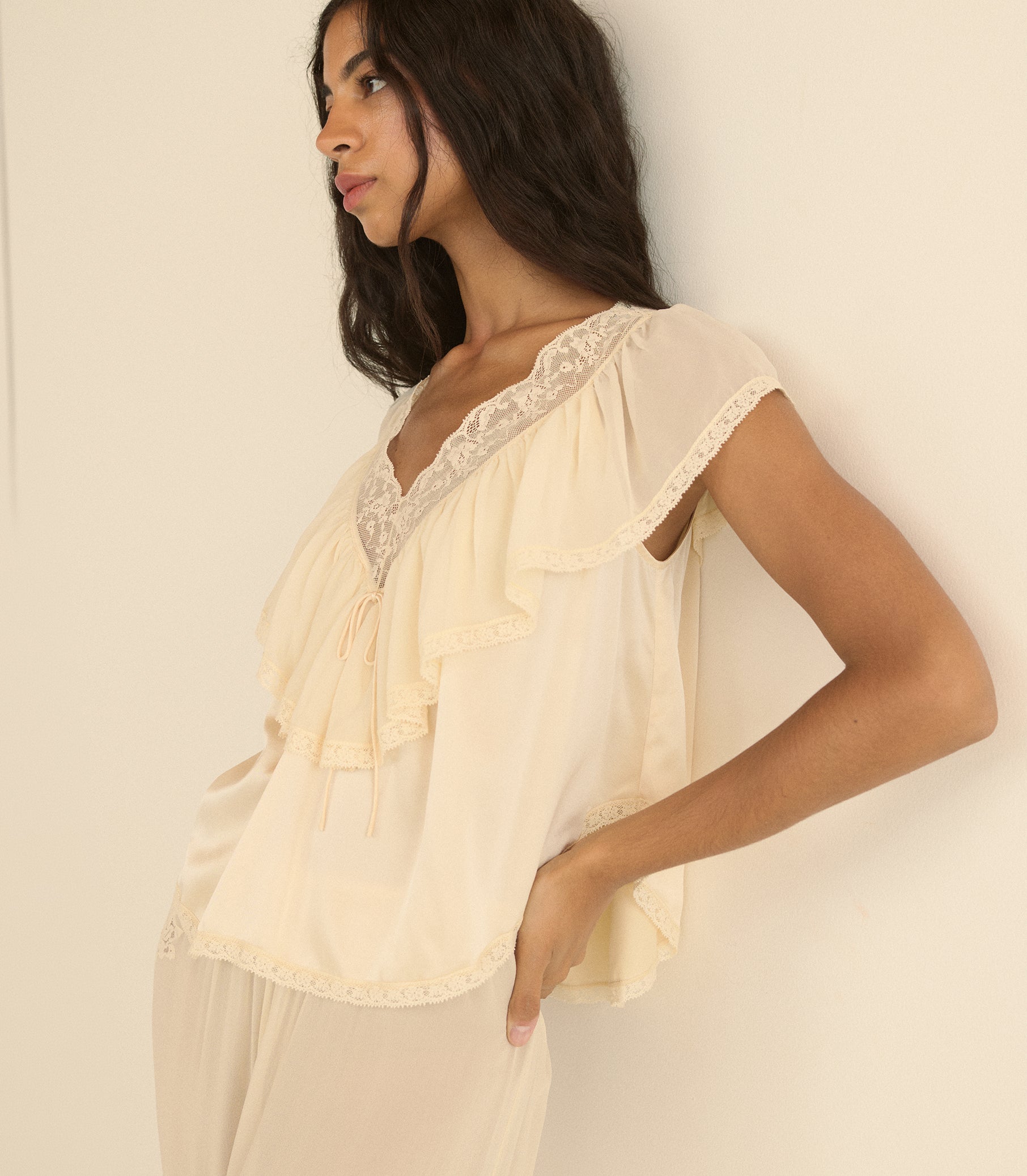 A woman wearing a cream-colored ruffled blouse with lace trim and matching pants, posing against a plain off-white wall.