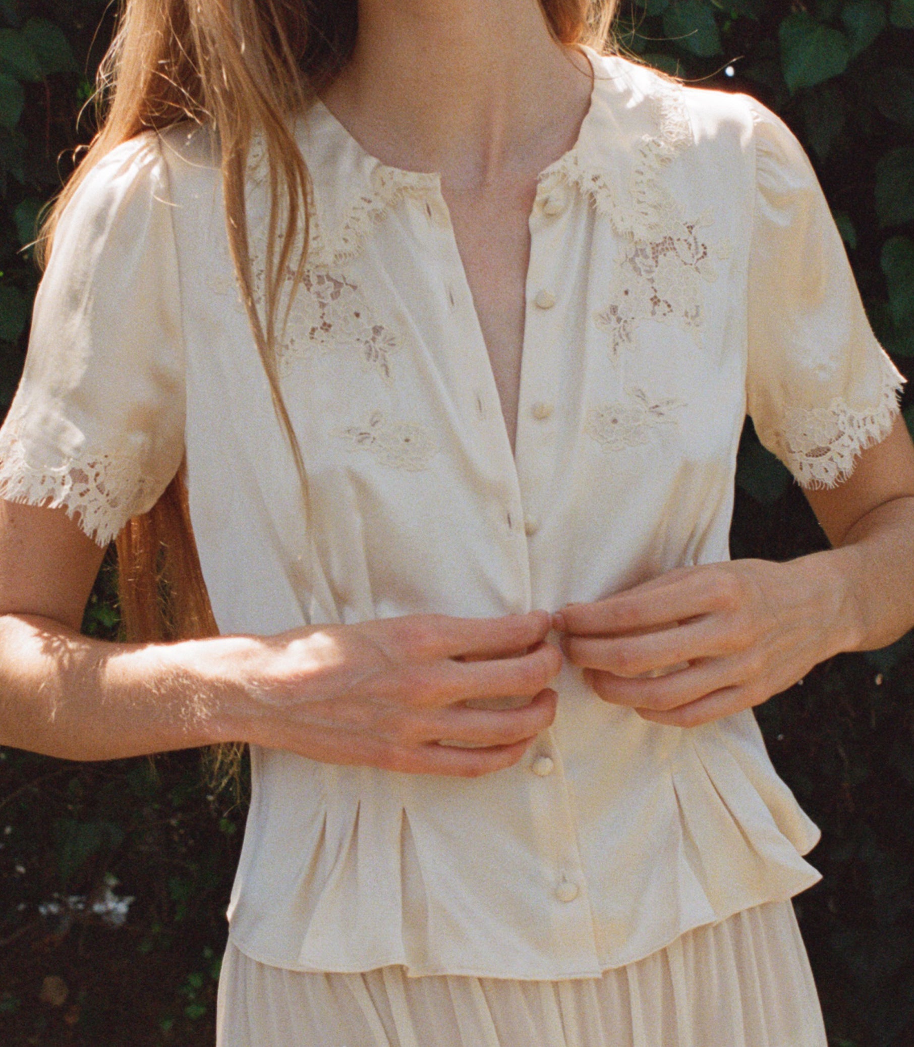 A woman wearing a cream-colored short-sleeve blouse with delicate floral embroidery and lace trim, buttoned up to the chest, set against a dark, leafy outdoor background.