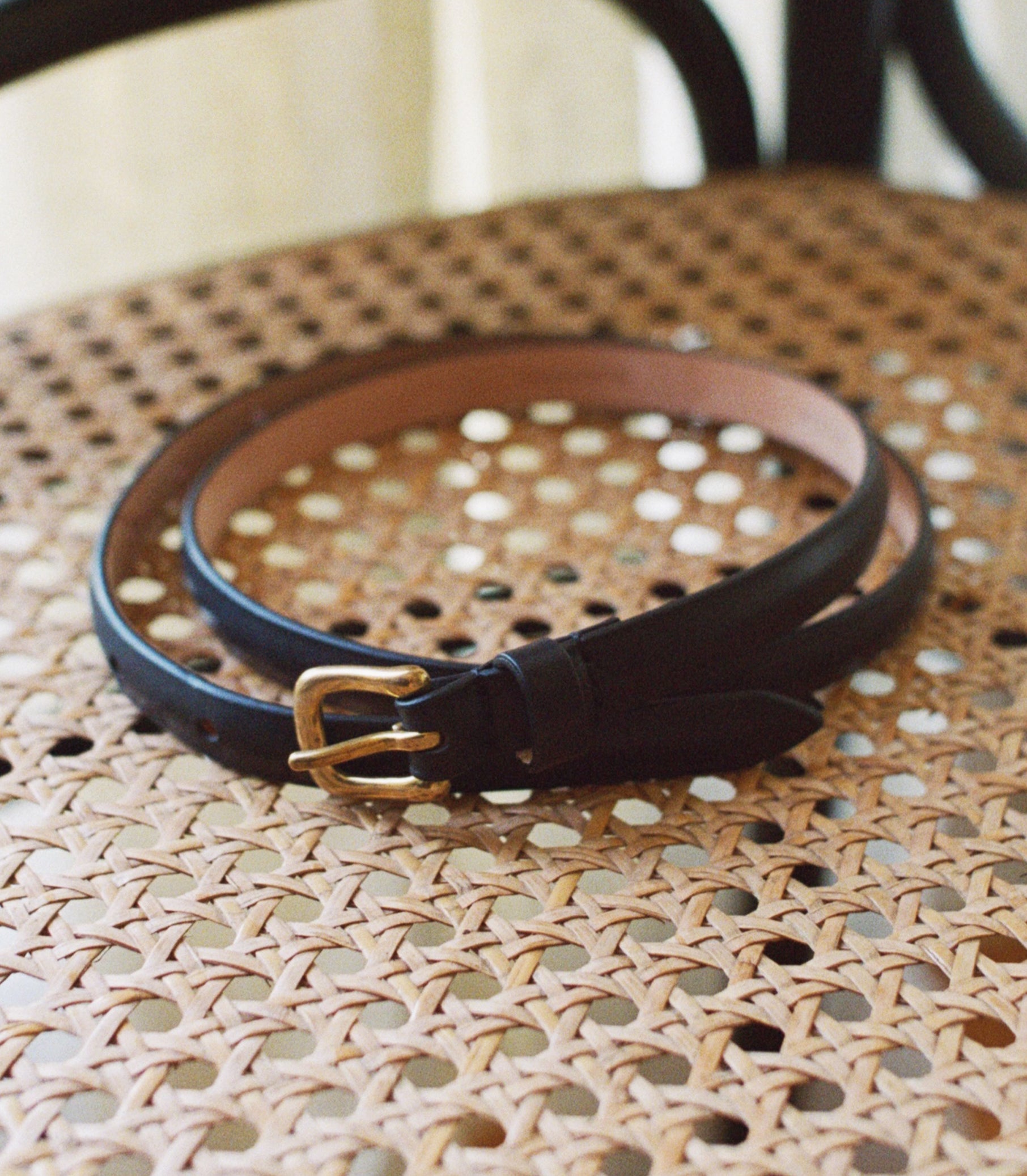 A black leather belt with a gold-tone buckle, coiled on a woven rattan chair seat. 1
