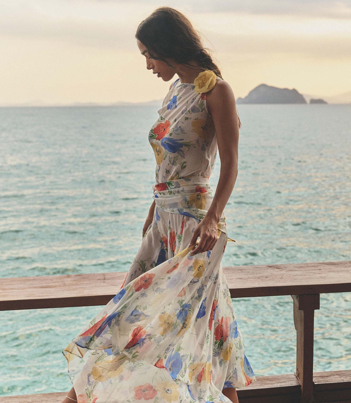 Woman in a white floral dress standing on a wooden platform by the ocean with mountains in the background.