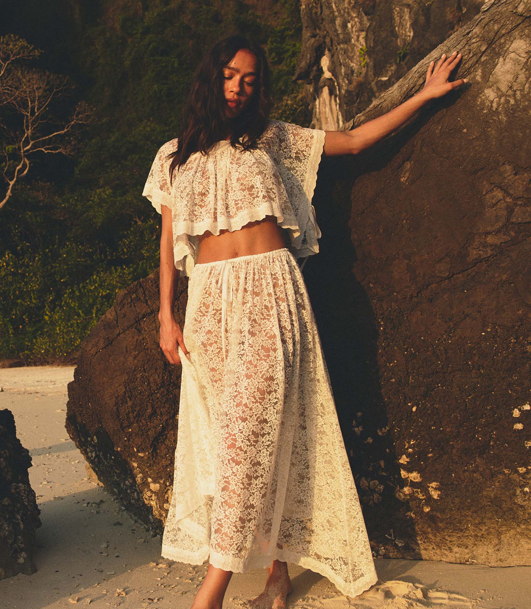 Woman in a white lace top and matching skirt standing against a rock with a natural background 1

